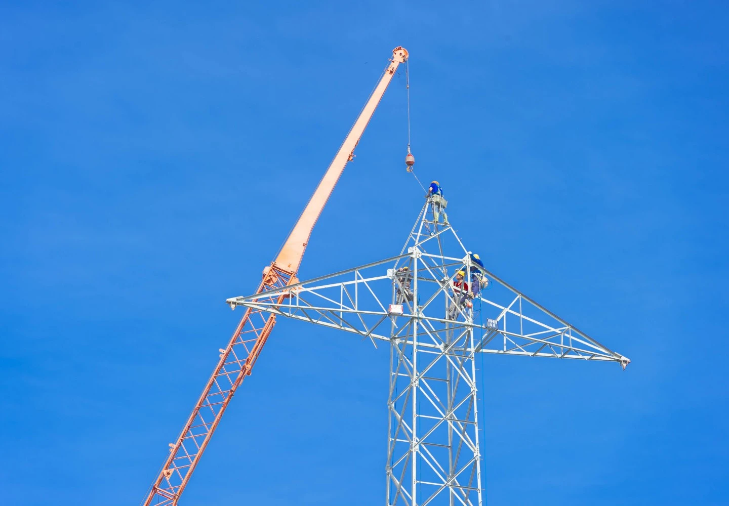 Workers in safety gear assemble or maintain a high-voltage steel lattice transmission tower using a crane against a clear blue sky.